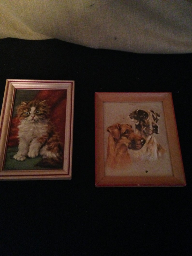 May include: Two framed vintage paintings. One shows a fluffy cat with brown and white fur, against a red background. The other depicts three dogs in shades of brown and tan.