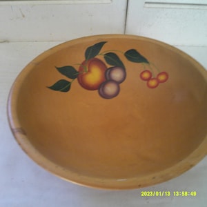Könnte beinhalten: Eine Holzschüssel mit handbemaltem Fruchtmuster. Die Schüssel ist hellbraun und die Früchte sind in Rot-, Gelb-, Lila- und Grüntönen bemalt.