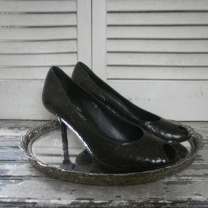 May include: A pair of black peep-toe high heels with a textured pattern. The shoes are displayed on a silver-colored, ornate tray. The background features white, slatted shutters.