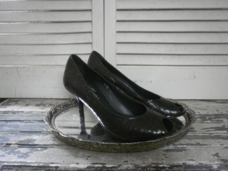 May include: A pair of black peep-toe high heels with a textured pattern. The shoes are displayed on a silver-colored, ornate tray. The background features white, slatted shutters.
