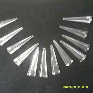 May include: A collection of 12 clear crystal teardrop-shaped prisms, arranged in a semi-circle on a black surface.