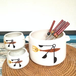 Op de afbeelding: Witte keramische fonduepot met een bruin en zwart ontwerp van een gestileerde lepel en vork. De pot staat op een geweven placemat met zes houten fonduevorken met rode handvatten.