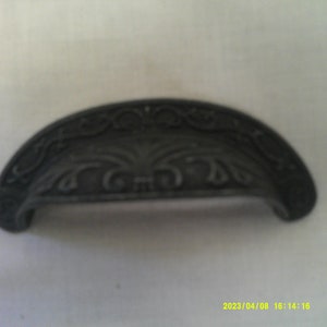 May include: A black metal drawer pull with an ornate design. The pull is shaped like an arch with a decorative pattern on the top.