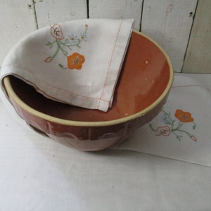 Puede incluir: Un tazón grande de cerámica marrón con un borde blanco y un diseño floral. El tazón está sentado sobre un mantel blanco con un diseño floral a juego.