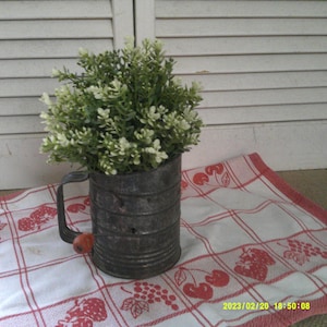 Może przedstawiać: Stary metalowy kubek miarowy z czerwoną rączką, wypełniony małym białym i zielonym krzewem, stojący na czerwono-białym wzorzystym obrusie.