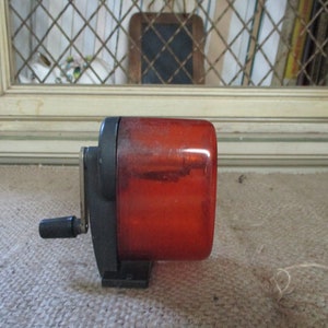 May include: A red plastic pencil sharpener with a black crank handle. The sharpener is sitting on a brown surface.