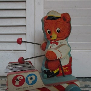 Könnte beinhalten: Vintage Holzspielzeug Xylophon mit roten Schlägeln. Das Xylophon hat ein rotes und blaues Fisher-Price Logo und einen Comic-Bären, der Xylophon spielt. Der Bär trägt ein weißes Hemd und einen blauen Hut. Das Spielzeug hat die Aufschrift "Tiny Teddy" auf der Seite.