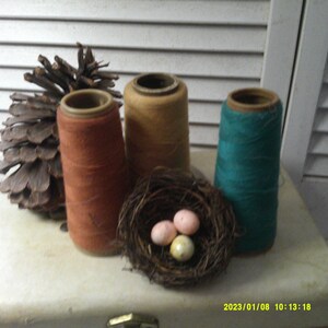 May include: Three spools of thread, one orange, one tan, and one teal, are arranged on a white surface with a pine cone and a small bird's nest with three speckled eggs.