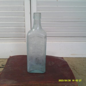 Puede incluir: Una botella de vidrio azul claro vintage con el texto en relieve "SCOTT'S EMULSION" en la parte delantera. La botella es rectangular con hombros redondeados y un cuello largo. Se encuentra sobre una superficie marrón oscuro.
