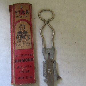 May include: Vintage 5 Star Handy Tool in a red and white box. The tool is made of metal and has a handle with a loop. The box has a picture of a woman using the tool to cut glass. The text on the box reads "5 Star Handy Tool", "Cuts Glass Like Diamond", "Will Last A Lifetime", and "Price $2.50".