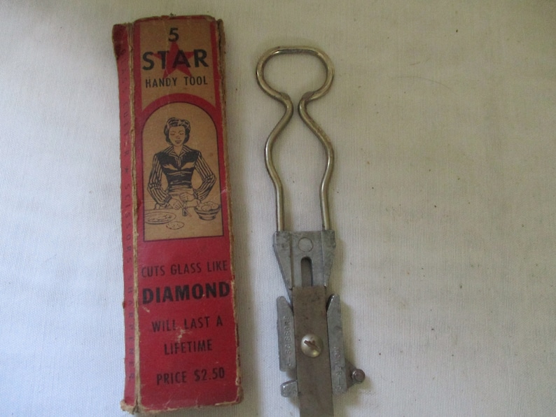 May include: Vintage 5 Star Handy Tool in a red and white box. The tool is made of metal and has a handle with a loop. The box has a picture of a woman using the tool to cut glass. The text on the box reads "5 Star Handy Tool", "Cuts Glass Like Diamond", "Will Last A Lifetime", and "Price $2.50".