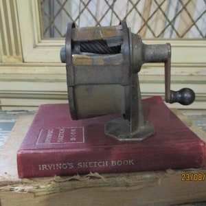 Pode incluir: Um apontador de lápis vintage de metal com manivela e base de metal. O apontador está em cima de um livro vermelho com o título "Irving's Sketch Book".