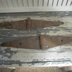 May include: Two rusty metal hinges, each with a triangular shape and a central hinge. The hinges are on a weathered wooden surface.