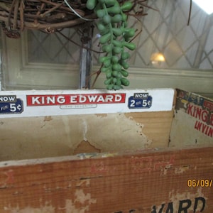 Könnte beinhalten: Eine antike Holzkiste mit einem Etikett, auf dem "King Edward The Seventh" und "Now 2 for 5c" steht. Die Kiste ist verwittert und hat eine verblichene braune Oberfläche. Die Kiste steht auf einem Tisch mit einer grünen Pflanze im Hintergrund.