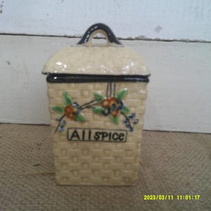 May include: A white ceramic spice jar with a black lid and a woven pattern. The jar has a floral design and the word "Allspice" written on the front.