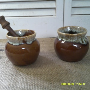 Può includere: Due vasi in ceramica smaltata marrone e bianca con un pestello di legno. I vasi hanno una smaltatura marrone screziata con accenti bianchi. Il pestello è in legno e ha una cima arrotondata.