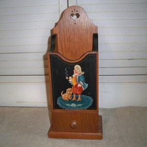 Könnte beinhalten: Eine hölzerne Wandbox mit einem bemalten Bild eines Mannes mit einem Hund. Der Mann trägt eine blaue Jacke, eine rote Hose und ein gelbes Hemd. Er hält eine Pfeife in der Hand. Der Hund ist braun und weiß. Die Box hat eine kleine Schublade unten.