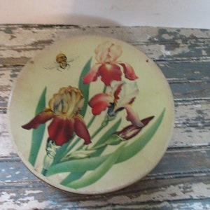 May include: A round, cream-colored tin with a floral design featuring three red, white, and yellow irises and a bee. The tin has a green background with green leaves.