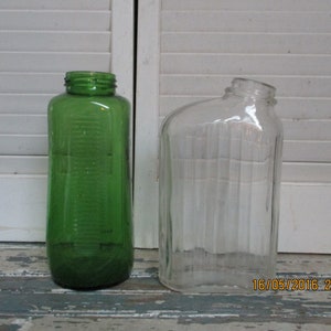 Op de afbeelding: Twee glazen flessen, een groene en een heldere, met een getextureerd oppervlak. Beide flessen zijn hoog en rechthoekig.