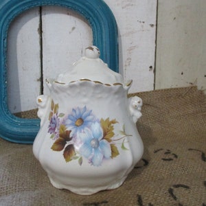 Op de afbeelding: Witte suikerpot van keramiek met een deksel en een bloemenmotief. De pot heeft een gouden rand en blauwe en bruine bloemen.