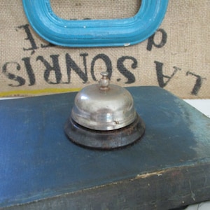 Pode incluir: Um sino de prata com uma base preta está sobre uma superfície azul. O sino é um sino de serviço tradicional, frequentemente usado em hotéis ou lojas.