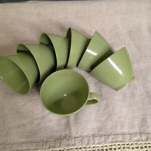 May include: A set of eight green ceramic cups arranged in a semi-circle on a beige linen tablecloth with a crocheted edge.