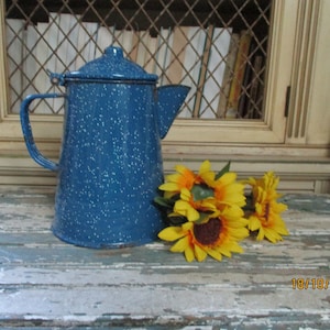Peut inclure: Une cafetière en émail bleu avec des mouchetures blanches repose sur une surface en bois vieilli à côté d'un bouquet de tournesols jaunes. La date 10/10/2016 est écrite sur la surface.