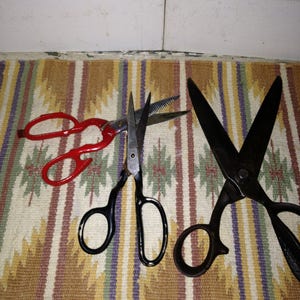 May include: Three pairs of scissors are displayed on a patterned rug. One pair is red, and the other two are black. The scissors vary in size and style, with one having a serrated blade. The rug has a geometric design with muted colors.