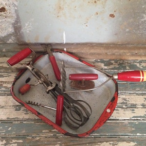 Peut inclure: Un bol mélangeur en émail rouge contenant une sélection d'ustensiles de cuisine vintage, dont un fouet, un batteur à œufs à manivelle, un ouvre-bouteille et un tire-bouchon. Les outils ont des poignées en plastique rouge et sont disposés soigneusement dans le bol.