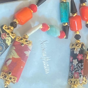 May include: A colorful necklace with a variety of beads and decorative elements. The necklace features cylindrical, rectangular, and spherical beads in shades of orange, red, blue, green, and purple. Some beads have floral patterns and gold accents. The necklace is on a white bag.