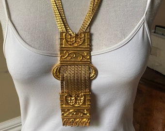 May include: A gold-toned statement necklace with a large, ornate pendant featuring intricate floral designs. The pendant is suspended from a chain of multiple strands.