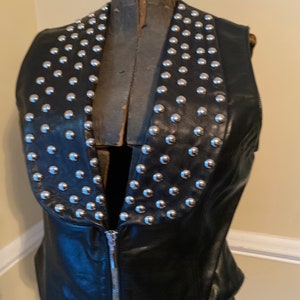 May include: A black leather vest with a silver studded collar. The vest has a zipper closure.