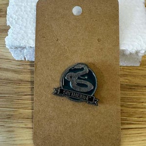 May include: A circular metal pin featuring a dark green background with a silver-toned snake emblem. Below the snake, a silver banner displays the text SLYTHERIN in black capital letters. The pin is attached to a rectangular brown card.
