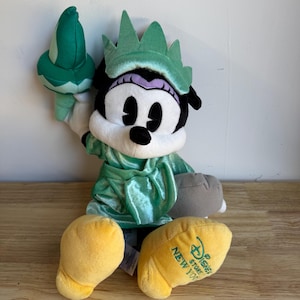 May include: A plush Minnie Mouse dressed as the Statue of Liberty. The doll has a green dress and crown, holding a green torch. The yellow feet have the text "Disney Store New York". The doll is sitting on a wooden surface.