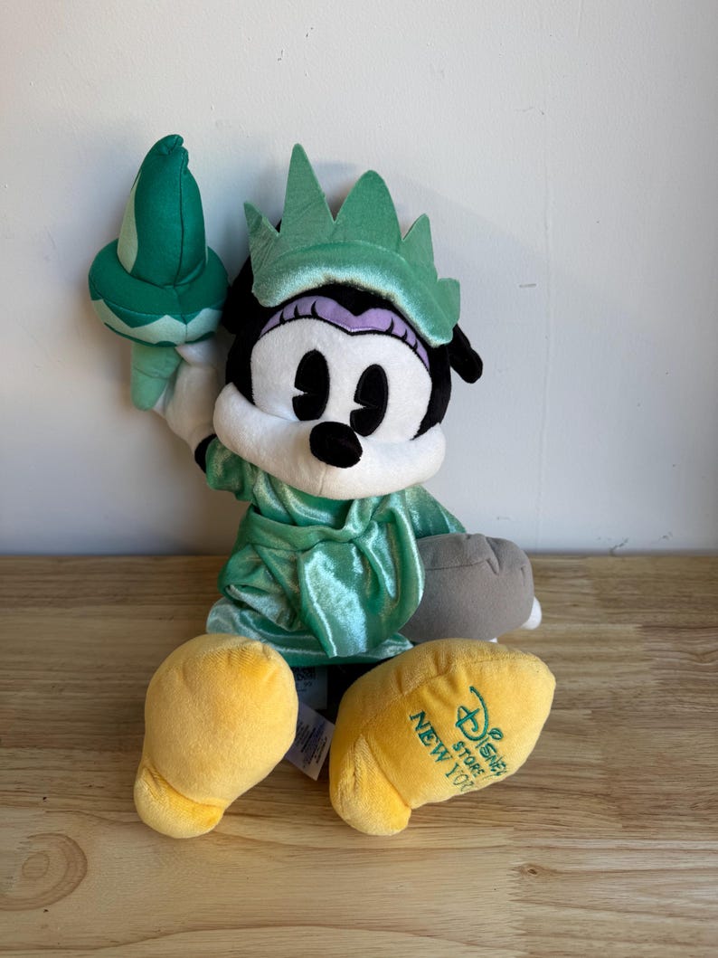 May include: A plush Minnie Mouse dressed as the Statue of Liberty. The doll has a green dress and crown, holding a green torch. The yellow feet have the text "Disney Store New York". The doll is sitting on a wooden surface.