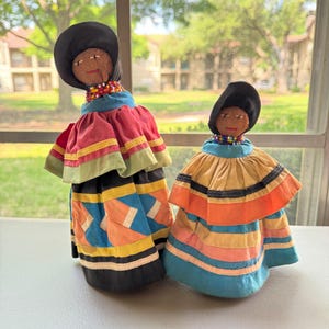 May include: Two handmade dolls with black hats and beaded necklaces. The dolls wear colorful patchwork dresses in shades of red, blue, yellow, and orange. The dolls are standing on a white surface in front of a window.