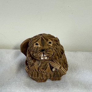 May include: A detailed, hand-carved wooden beaver figurine. The beaver is brown with visible teeth and is holding a small log. The carving has textured fur and is set on a white surface.