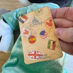 May include: A plush toy with a black head and white face, wearing a teal outfit. A Disney-branded tag features stamps with flags of various countries. The tag reads "Plush Peluche".
