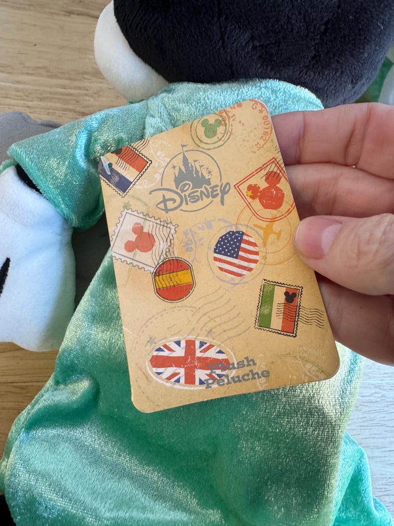 May include: A plush toy with a black head and white face, wearing a teal outfit. A Disney-branded tag features stamps with flags of various countries. The tag reads "Plush Peluche".