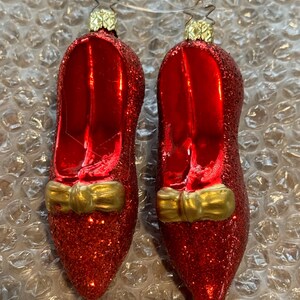 May include: A pair of red glass slipper Christmas ornaments with gold bows. The ornaments are covered in glitter.