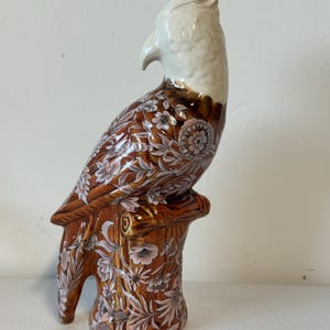 Puede incluir: Figura de loro de cer&aacute;mica con cabeza blanca y cuerpo marr&oacute;n adornado con motivos florales. El loro est&aacute; posado sobre una base de toc&oacute;n de &aacute;rbol marr&oacute;n. La figura tiene un acabado brillante y es probablemente un art&iacute;culo decorativo.