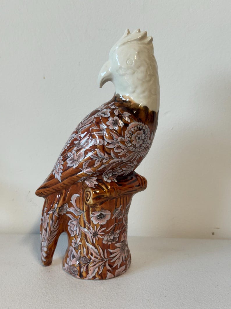 Puede incluir: Figura de loro de cer&aacute;mica con cabeza blanca y cuerpo marr&oacute;n adornado con motivos florales. El loro est&aacute; posado sobre una base de toc&oacute;n de &aacute;rbol marr&oacute;n. La figura tiene un acabado brillante y es probablemente un art&iacute;culo decorativo.