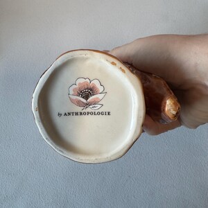 Puede incluir: Una taza de cer&aacute;mica marr&oacute;n con asa, vista desde abajo. La base interior es de color crema con un dise&ntilde;o floral rosa y negro y el texto "by ANTHROPOLOGIE". El exterior de la taza tiene un aspecto texturizado similar a la corteza.