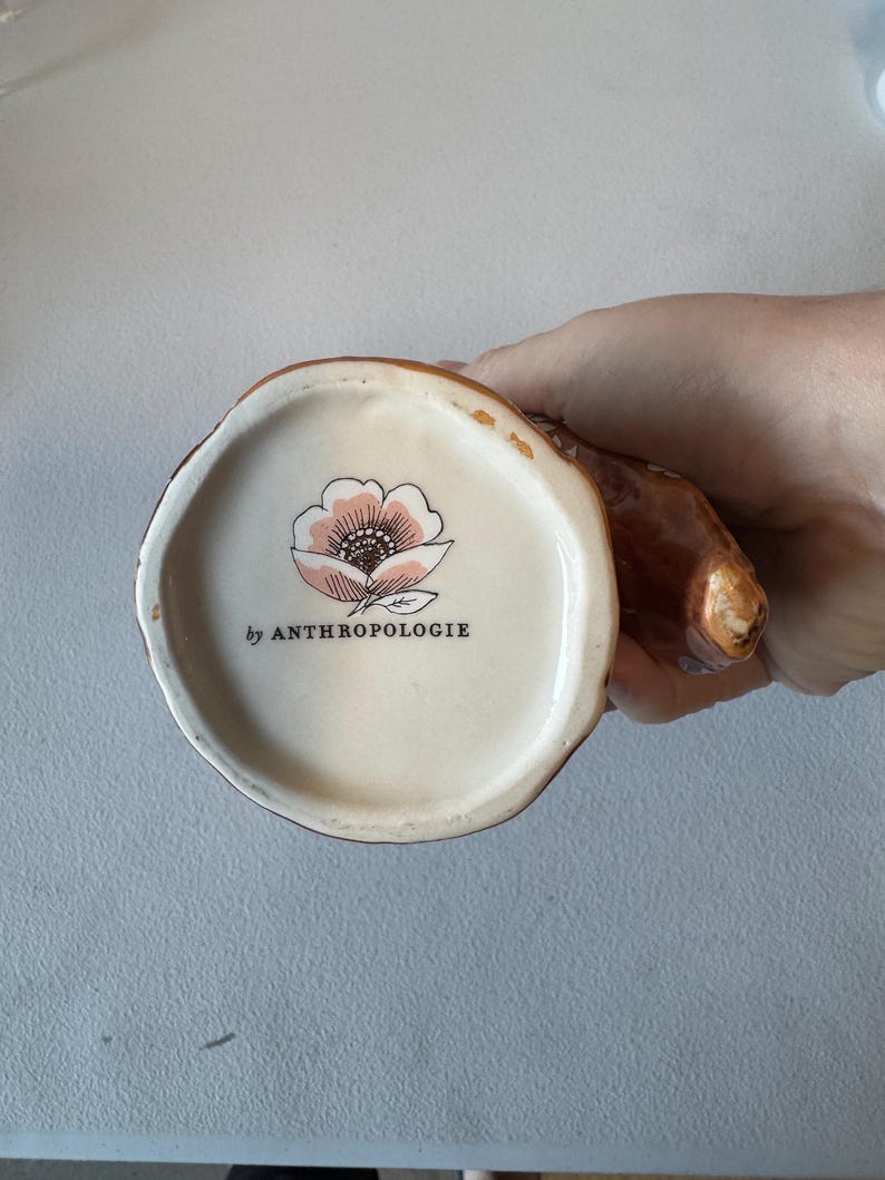 Puede incluir: Una taza de cer&aacute;mica marr&oacute;n con asa, vista desde abajo. La base interior es de color crema con un dise&ntilde;o floral rosa y negro y el texto "by ANTHROPOLOGIE". El exterior de la taza tiene un aspecto texturizado similar a la corteza.
