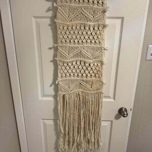 May include: A cream-colored macrame wall hanging featuring intricate knotted patterns, including geometric chevron shapes and textured square knots, finished with a long, loose fringe at the bottom, suspended from a black plastic clothes hanger.