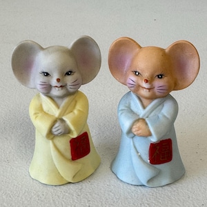 May include: Two ceramic mouse figurines. One mouse wears a yellow robe, the other a blue robe. Both have large ears, smiling faces, and red patches with Chinese characters. The figurines are decorative and likely part of a collection.