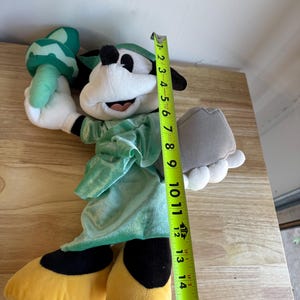 May include: A plush toy of a cartoon character dressed as the Statue of Liberty. The toy is white, black, yellow, and green, and measures approximately 15 inches tall. It holds a torch and a tablet.