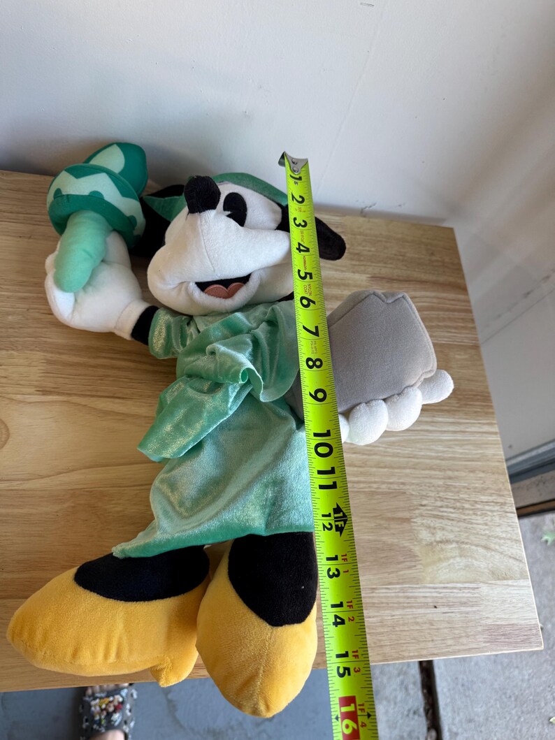 May include: A plush toy of a cartoon character dressed as the Statue of Liberty. The toy is white, black, yellow, and green, and measures approximately 15 inches tall. It holds a torch and a tablet.