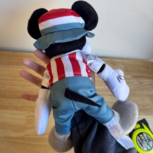 Pu&ograve; includere: Un pupazzo di peluche di Topolino con una camicia a righe rosse e bianche, pantaloni blu e un cappello abbinato. La bambola ha orecchie nere, guanti bianchi e scarpe grigie. La bambola &egrave; tenuta da una persona.