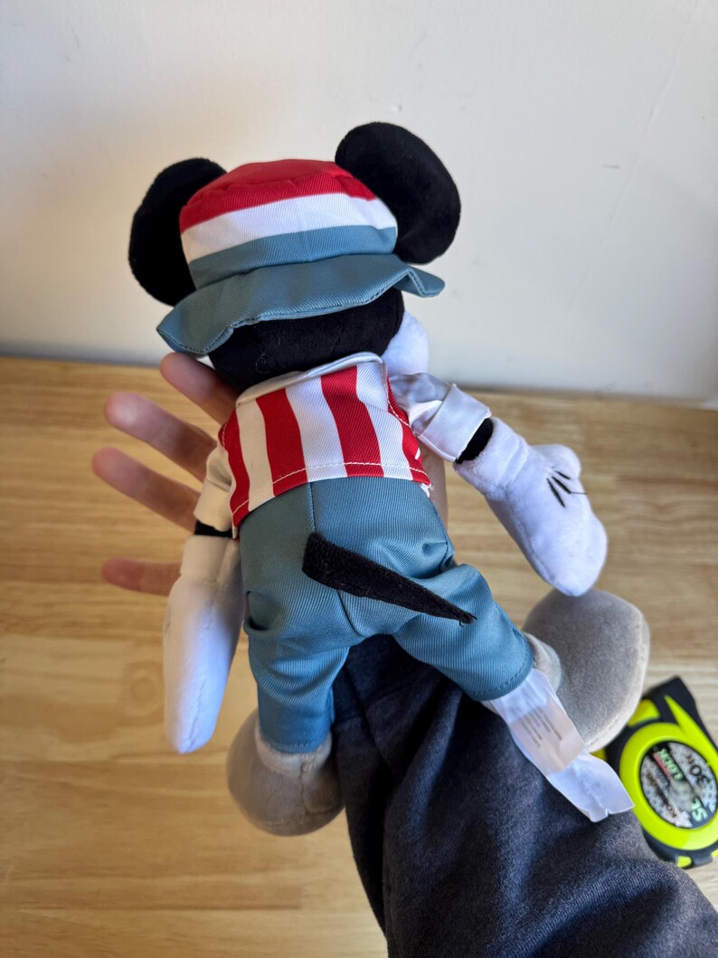 Pu&ograve; includere: Un pupazzo di peluche di Topolino con una camicia a righe rosse e bianche, pantaloni blu e un cappello abbinato. La bambola ha orecchie nere, guanti bianchi e scarpe grigie. La bambola &egrave; tenuta da una persona.